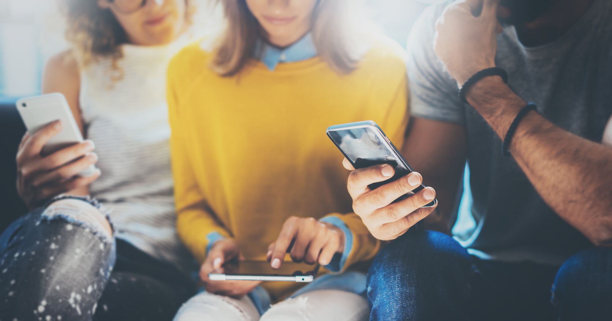 Three young adults sit together, each focused on their smartphones. One wears a yellow sweater, another holds a tablet, highlighting modern digital interaction.