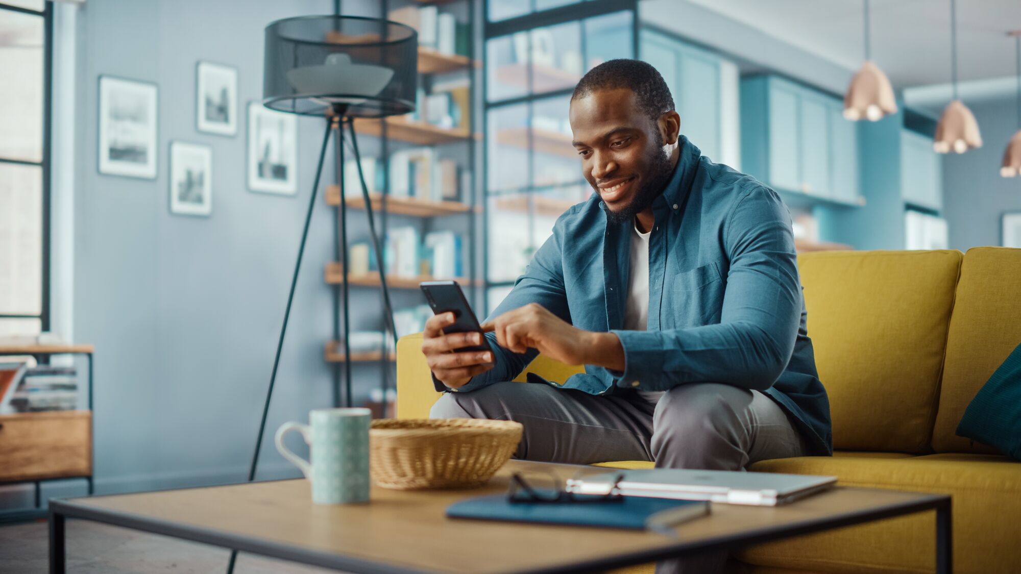 Man sitting on a yellow couch, smiling while using his smartphone. A coffee mug and a basket sit on the coffee table in a modern living room.